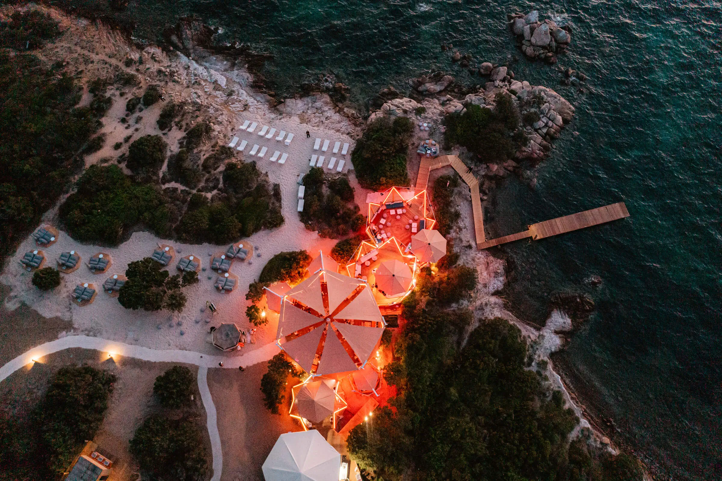 Aerial view of a coastal beach resort with illuminated lounge tents, sunbeds, and a wooden pier by the sea.