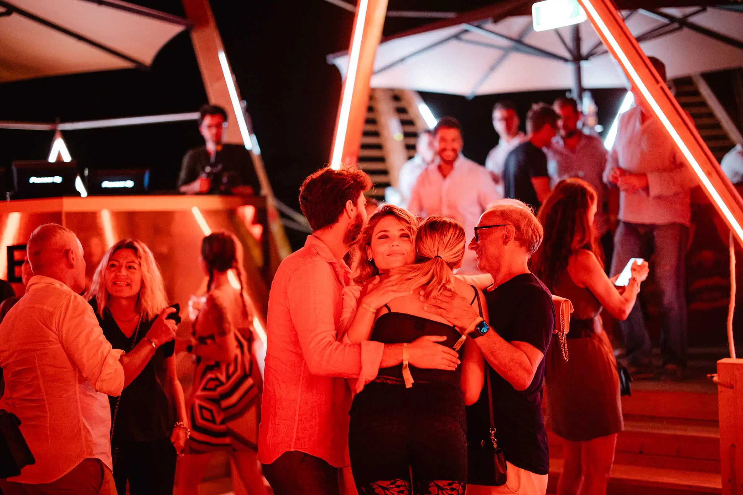 Guests socializing and hugging at a lively outdoor evening event at Cone Club, with red neon lighting and a DJ booth in the background.